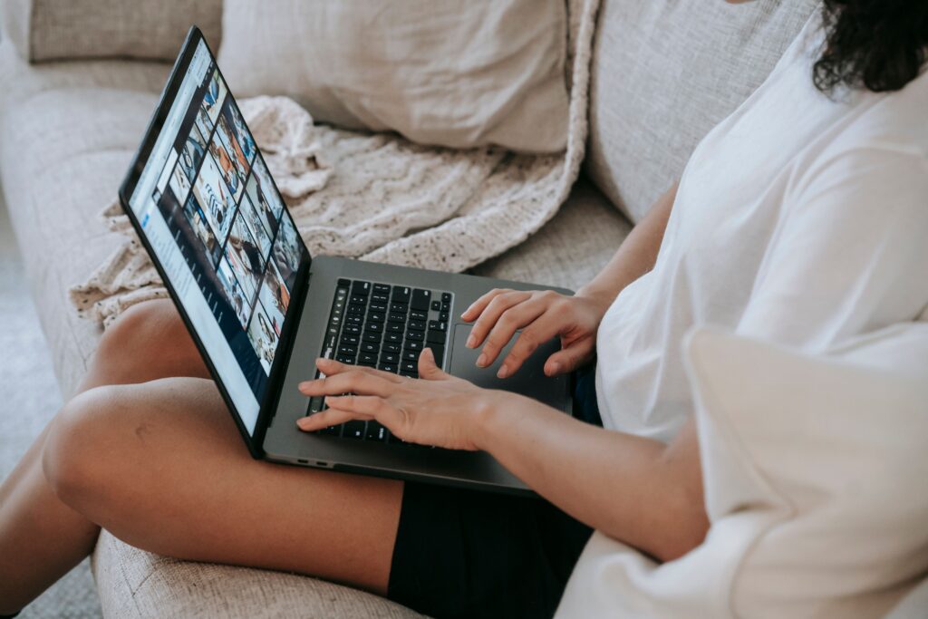 From above side view of crop faceless woman in casual clothes relaxing on comfortable sofa and watching photos on laptop during weekend at home