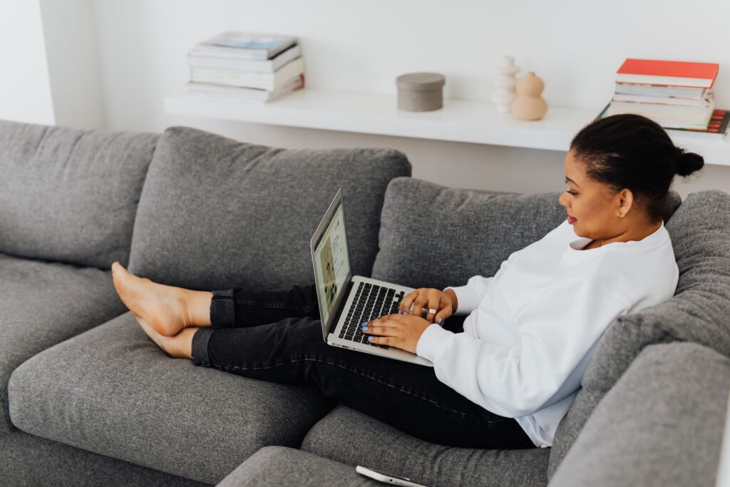 Woman in white sweater using laptop while lying on gray sofa indoors.